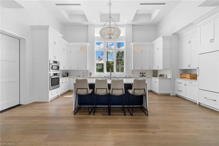 Kitchen with a high ceiling, white cabinets, an island with sink, light stone counters, and glass insert cabinets