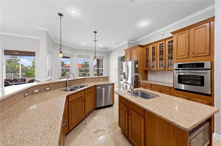 Kitchen featuring brown cabinets, light stone counters, stainless steel appliances, glass insert cabinets, and a center island with sink