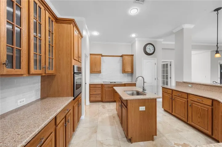 Kitchen with decorative backsplash, light stone counters, decorative light fixtures, a center island with sink, and crown molding