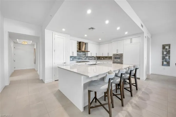 Kitchen featuring a breakfast bar area, a center island with sink, white cabinets, wall chimney range hood, and backsplash