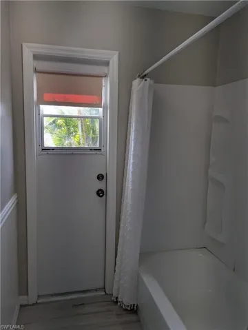 Bathroom featuring wood-finish flooring, white wainscoting, a full tub with shower, and a windowed exterior door