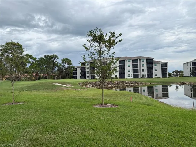 Multi-story residential building featuring screened balconies, situated on a lot with expansive lawn, mature landscaping, and a water feature