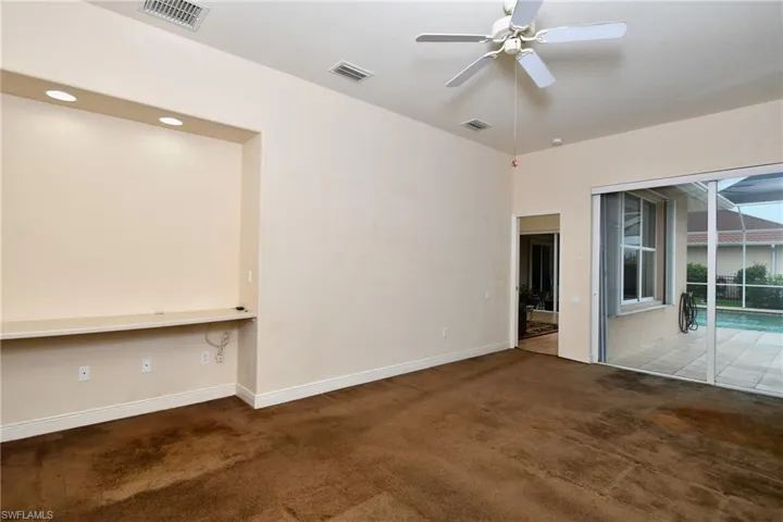 Carpeted interior space featuring a built-in media niche with integrated lighting, a ceiling fan, and sliding glass doors opening to a screened patio with a pool