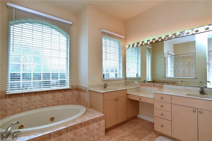 Bathroom featuring a large soaking tub with tiled surround, dual vanity sinks, extensive counter space, and a full-width mirror with integrated lighting