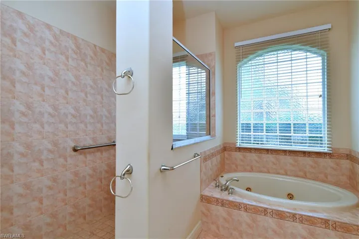 Bathroom featuring a jetted soaking tub with tiled surround, a separate shower with grab bar, an arched window with blinds, and mirrored wall