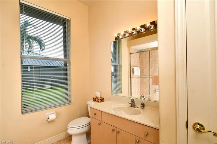Full bathroom featuring a single window with blinds, a vanity with an integrated sink and light-toned countertop, wood-finish cabinetry with silver-tone hardware, and a mirrored vanity light bar