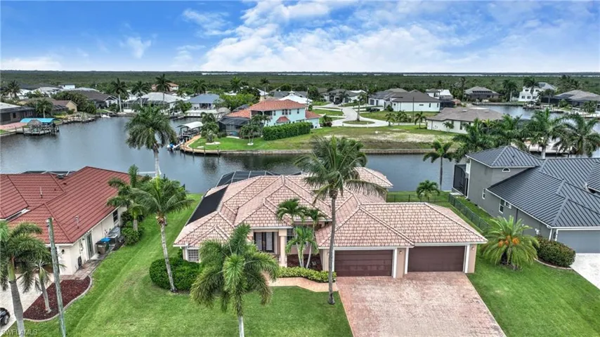 Waterfront residence featuring a tile roof, dual garage doors, a paver driveway, lush green landscaping, and mature palm trees
