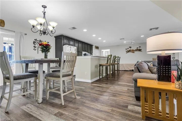 Open-concept living area featuring wood-finish flooring, a dining area with a chandelier, a kitchen island with seating, dark cabinetry, and white walls