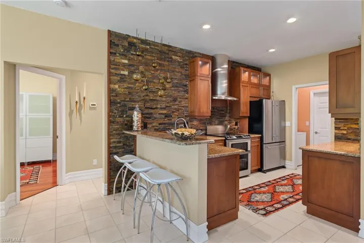 Kitchen featuring light tile patterned flooring, tasteful backsplash, wall chimney range hood, light stone countertops, and appliances with stainless steel finishes