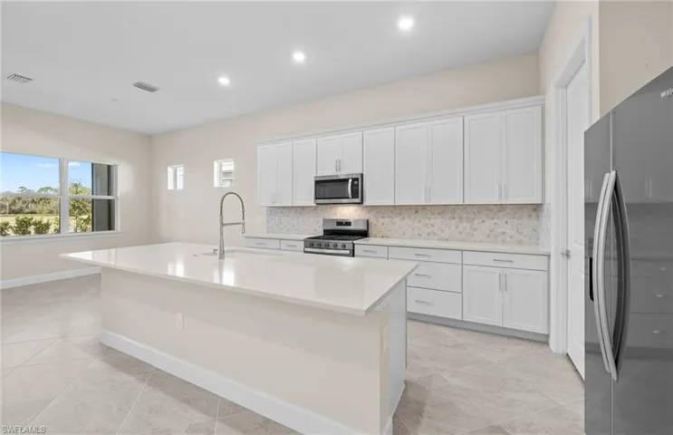 Kitchen featuring backsplash, stainless steel appliances, white cabinets, a center island with sink, and light stone countertops