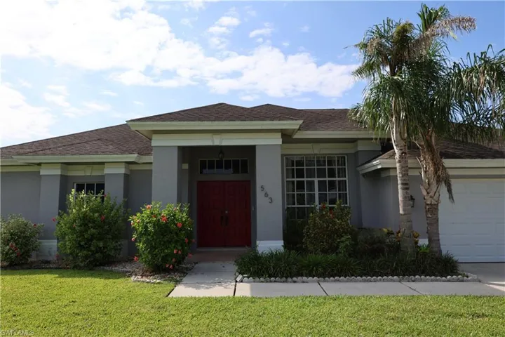 Light gray stucco exterior featuring a red double-door entry with transom window, white trim accents, a dark shingle roof, established landscaping, and a concrete driveway