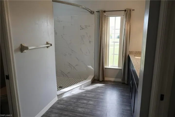Bathroom featuring a walk-in shower with white marble-patterned tile walls and black and white penny tile floor, wood-finish flooring, a double vanity with a light-colored countertop, and a window with natural light