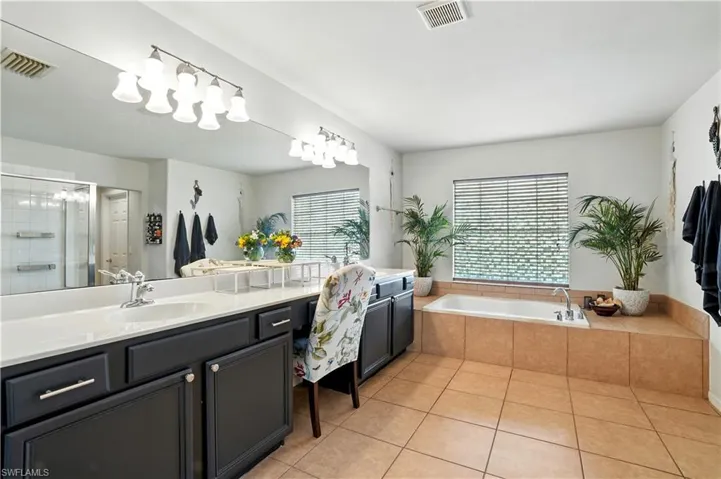 Luxurious primary bathroom with dual vanities, abundant counter space, and elegant dark cabinetry. A large soaking tub beneath a bright window offers a serene retreat, while the separate walk-in shower adds convenience. Warm tile flooring and stylish lighting create a spa-like ambiance, perfect for unwinding after a long day. This spacious en-suite blends comfort and function, making it an inviting highlight of this beautiful Southwest Florida home