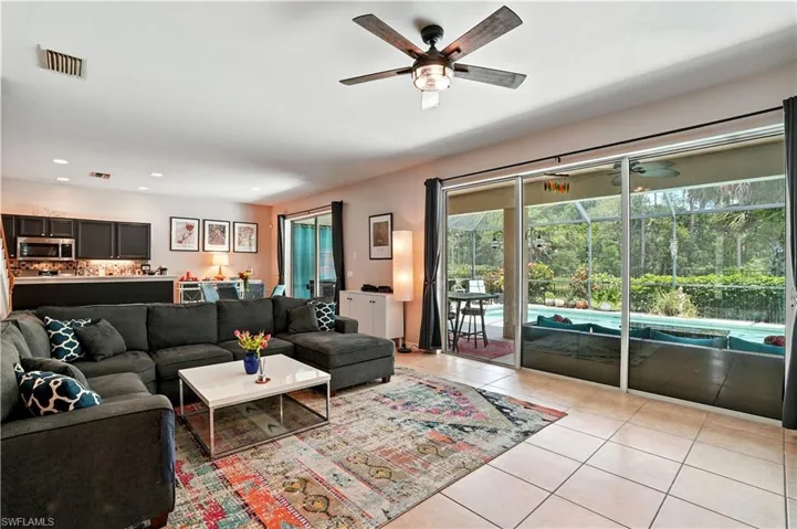 Bright and inviting open-concept living room with tile floors, recessed lighting, and a modern ceiling fan. Large sliding glass doors showcase serene pool and preserve views, filling the space with natural light. Seamless flow to the dining area and updated kitchen with dark cabinetry and stylish backsplash makes entertaining effortless. Perfect for Florida living, this spacious layout connects indoor comfort with outdoor enjoyment—ideal for families or seasonal retreats near Gulf beaches.