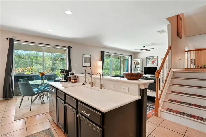 Modern kitchen with sleek dark cabinetry, stainless steel appliances, and a stylish mosaic tile backsplash. A spacious breakfast bar with seating opens to the dining area and living room, creating a seamless flow for entertaining. Large sliding glass doors provide abundant natural light and direct access to the screened pool and patio. Perfect for Florida living, this functional and inviting space combines contemporary design with comfort—ideal for gatherings near Gulf Coast attractions.