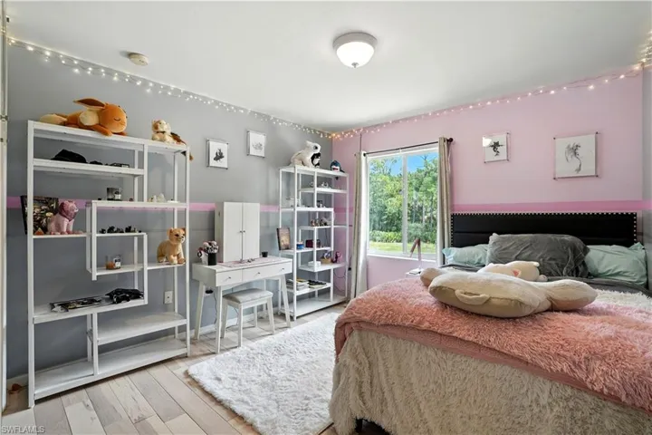 Charming bedroom with soft pink and gray accents, wood flooring, and a large window overlooking lush greenery. String lights add a cozy, playful touch, while open shelving and a vanity provide functional storage and style. Perfect as a guest room, child’s room, or creative space, this inviting retreat blends comfort and personality