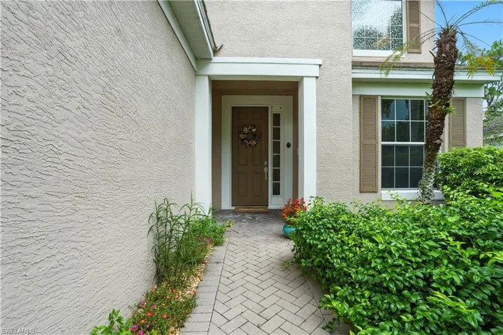 Inviting front entry with a charming covered porch, paver walkway, and vibrant tropical landscaping. The neutral-toned exterior, decorative shutters, and full-light side panel create a warm, welcoming first impression. Perfect curb appeal for this Southwest Florida home, blending style and privacy in a desirable community close to Gulf beaches and amenities.