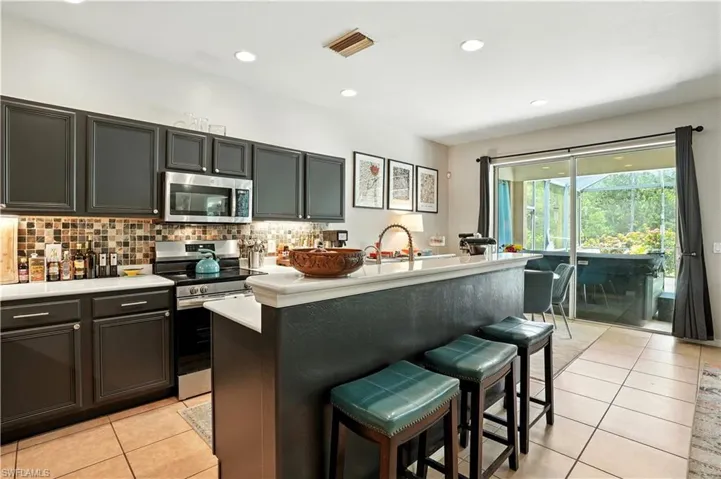 Modern kitchen with sleek dark cabinetry, stainless steel appliances, and a stylish mosaic tile backsplash. A spacious breakfast bar with seating opens to the dining area and living room, creating a seamless flow for entertaining. Large sliding glass doors provide abundant natural light and direct access to the screened pool and patio. Perfect for Florida living, this functional and inviting space combines contemporary design with comfort—ideal for gatherings near Gulf Coast attractions.