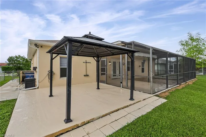 Expansive concrete patio with a black metal gazebo, leading to a screened-in pool area featuring a covered lanai and a concrete pool deck