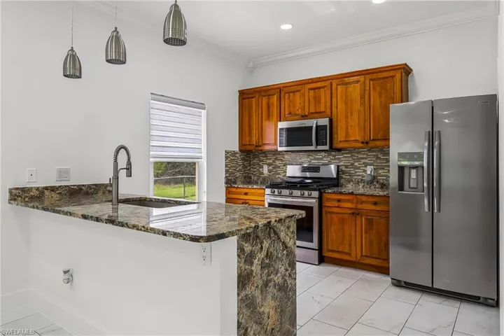 Kitchen featuring stone countertops, stainless steel appliances, wood cabinetry, a tile backsplash, and pendant lighting