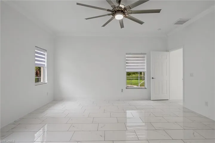 Spacious room featuring white tile flooring, white walls, two windows with blinds, a ceiling fan with integrated lighting, and crown molding
