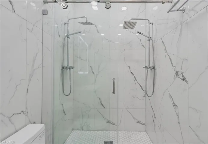 Shower enclosure featuring a glass door, dual rainfall showerheads, handheld shower wands, white marble-look tile, and a white hexagonal tile floor