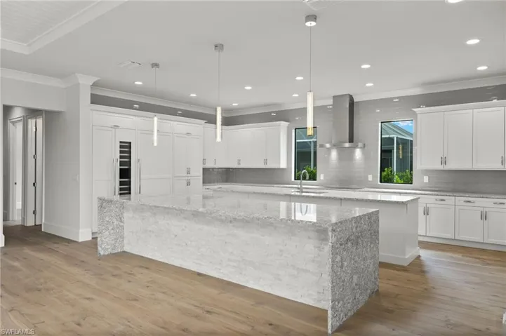 Kitchen featuring crown molding, light wood-type flooring, a spacious island, wall chimney exhaust hood, and light stone counters