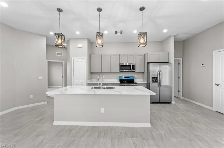 Kitchen featuring gray cabinets, stainless steel appliances, light stone counters, an island with sink, and pendant lighting
