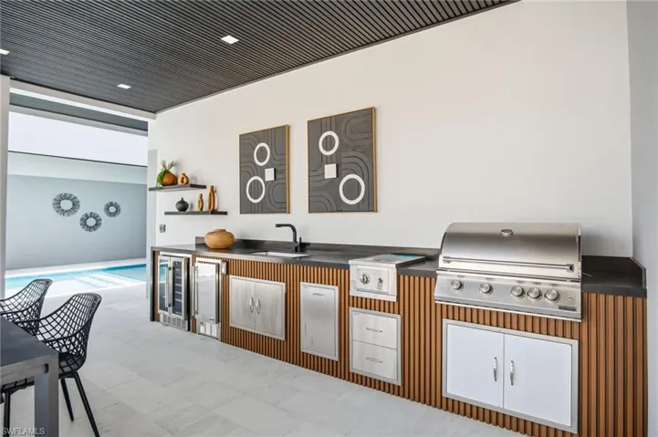 Outdoor kitchen featuring a built-in stainless steel grill, integrated beverage cooler, sink with a matte black faucet, and vertical wood-finish cabinetry panels