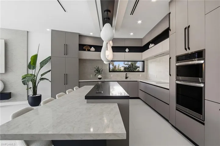 Contemporary kitchen featuring an oversized island with a light-toned countertop, sleek flat-panel cabinetry, integrated double wall ovens, a linear window, and recessed lighting