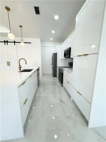 Kitchen with modern cabinets, white cabinetry, appliances with stainless steel finishes, decorative light fixtures, and light stone countertops