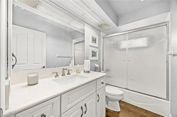 White vanity with recessed paneling and dark hardware, integrated rectangular sink, wall-mounted mirror, and wood-finish flooring