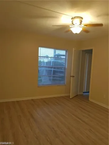 Wood-finish flooring throughout, featuring a ceiling fan with integrated lighting, a window with blinds, and a white interior door