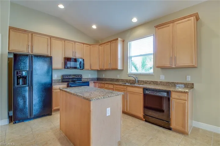 Kitchen featuring black appliances, light brown cabinetry, light stone counters, vaulted ceiling, and a center island