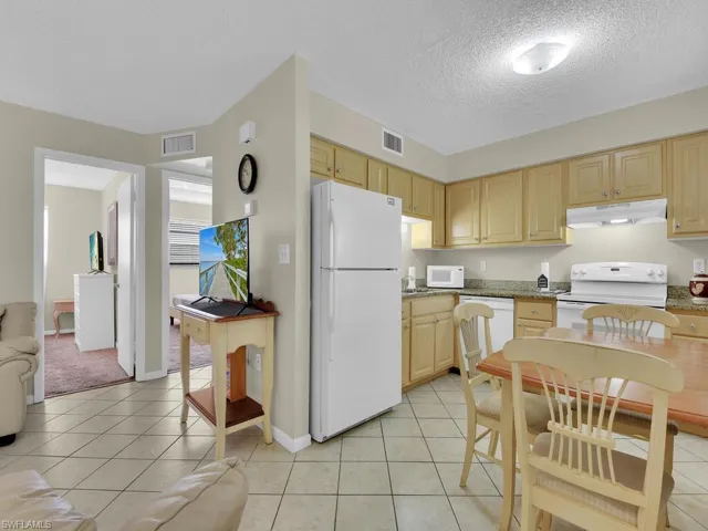 Kitchen with dark stone counters, light brown cabinetry, white appliances, light carpet, and a textured ceiling