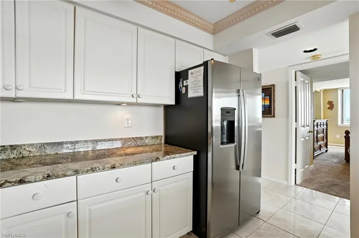 Kitchen featuring stainless steel refrigerator with ice dispenser, white cabinets, dark stone countertops, and light tile patterned flooring - Virtually Edited Image