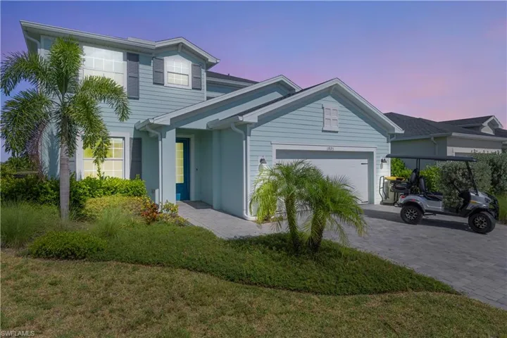 Two-story siding exterior with contrasting shutters, attached garage, paver driveway, and mature tropical landscaping
