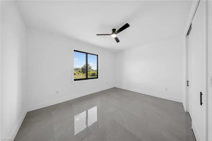 Spacious room featuring a single window with black trim, a ceiling fan with integrated lighting, white walls, and large format grey tile flooring