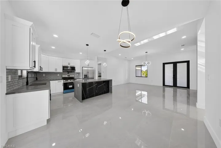 Open-concept kitchen and living area featuring a large central island with a marbled black finish, white shaker cabinetry, stainless steel appliances, and sleek gray tile flooring