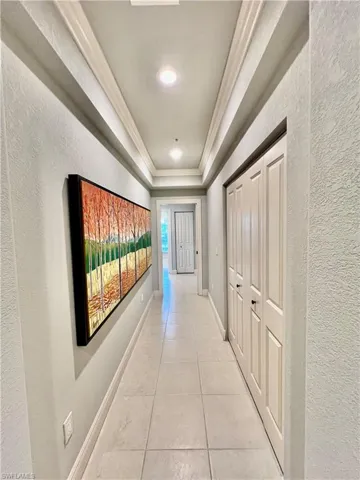 Corridor featuring baseboards, ornamental molding, light tile patterned floors, a textured wall, and a raised ceiling