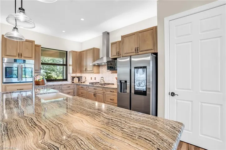 Kitchen with light stone countertops, wall chimney exhaust hood, appliances with stainless steel finishes, decorative light fixtures, and sink
