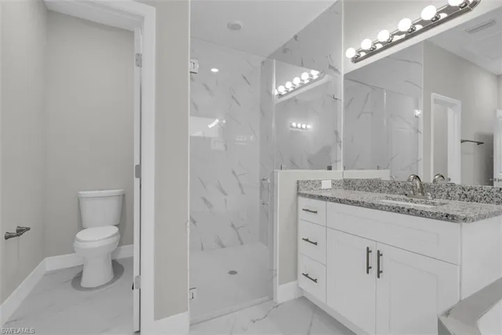 Bathroom featuring a clear glass shower enclosure with marble-patterned tile, a white vanity with a speckled countertop, an undermount sink, and a wall-mounted light fixture above a large mirror