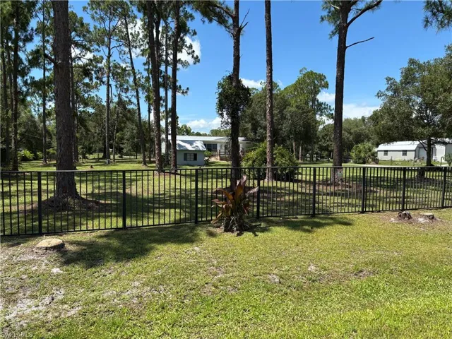 Expansive grassy area featuring a black metal fence, mature trees, and a distant white structure