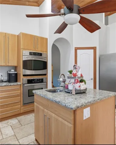 Kitchen with light stone countertops, stainless steel appliances, stone tile floors, an island with sink, and a ceiling fan
