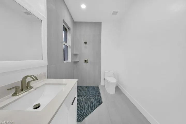 Modern vanity with a light-toned countertop, integrated rectangular sink, and metallic faucet, complemented by a walk-in shower featuring grey tiled walls, built-in shelving, and dark herringbone-patterned floor tiles