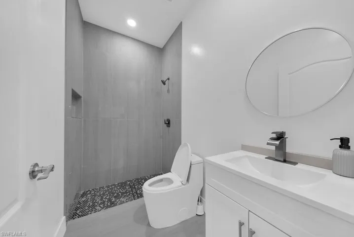 Bathroom featuring a walk-in shower with vertical tile surround and recessed shelving, pebble-style shower floor, oval wall-mounted mirror, and vanity with an integrated sink and chrome faucet
