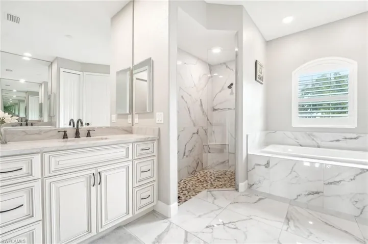 Spacious bathroom featuring a white vanity with dual sinks, matte black fixtures, a built-in soaking tub, a walk-in shower with pebble tile flooring, and large format marble-look tile throughout