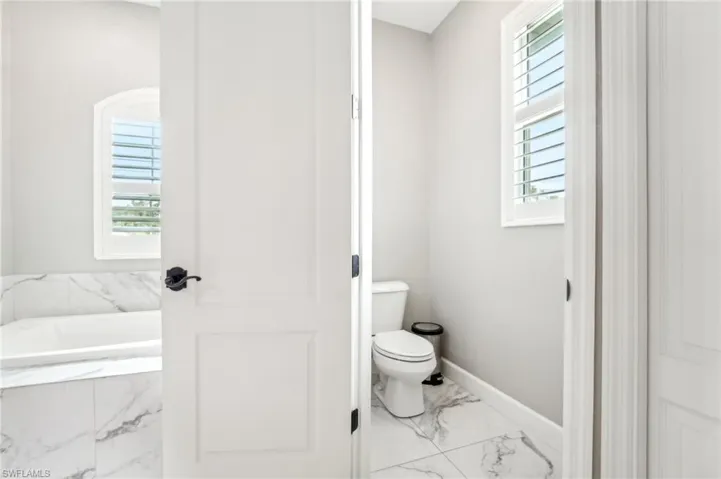 Bright bathroom interior featuring a built-in tub with marble-look surround, a private water closet with a window, white panel doors, and tile flooring