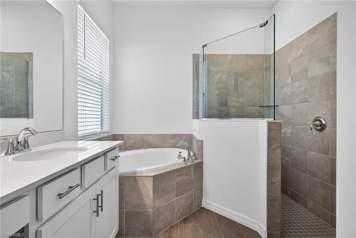 Primary Bathroom featuring a vanity with a white countertop and double sinks, a window with blinds, a corner bathtub, and a walk-in shower with tile walls and a glass partition