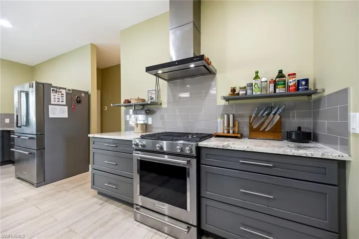 Kitchen with appliances with stainless steel finishes, ventilation hood, light stone countertops, gray cabinets, and backsplash